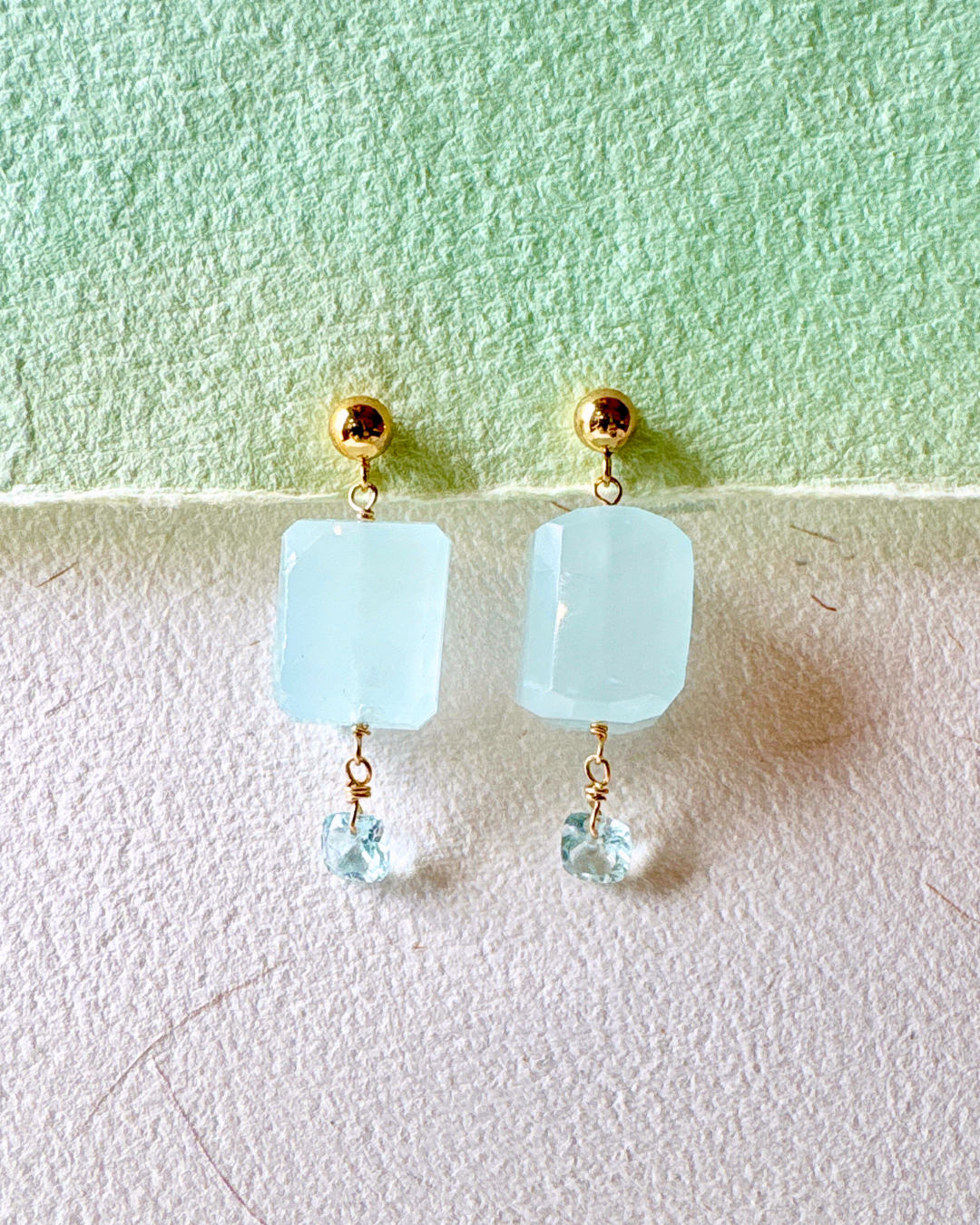 Pair of earrings with light blue square beads and gold accents on a light green and white background.